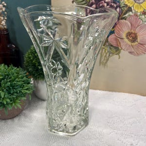 May include: Clear glass vase with a star and diamond pattern. The vase has a flared top and a hexagonal base. The vase is empty and sits on a white surface. The background includes a floral print and small green plants.