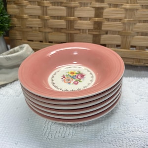 May include: A stack of six round ceramic bowls with a solid dusty pink rim and a cream-colored center. Each bowl features a central floral decal with yellow, pink, and blue flowers surrounded by a delicate black decorative border pattern.