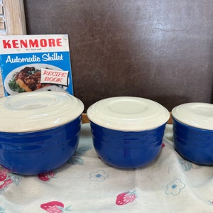 May include: Three vintage blue and white ceramic bowls with lids. The bowls are graduated in size and have a classic design. A recipe book titled "Kenmore Automatic Skillet" is in the background.