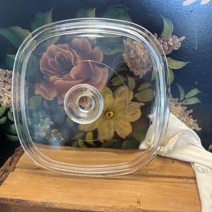 May include: A clear, square glass lid with a central knob. The lid has a slightly raised rim and is designed to fit a square dish. The background features floral artwork with roses and other flowers.