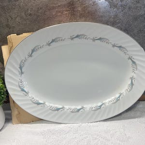May include: An oval white serving platter with a silver rim and a decorative border of blue and gray leaf-like designs. The platter is set on a white surface, suggesting a dining or serving context.