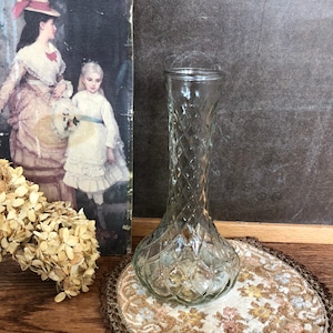 May include: A clear glass vase with a diamond pattern sits on a brown and white crocheted doily. The vase is tall and slender with a round base.