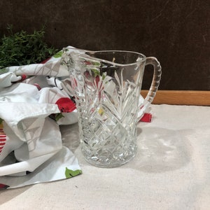 May include: A clear glass pitcher with a cut glass design. The pitcher has a handle and is sitting on a white fabric surface.