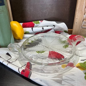 May include: A clear glass serving tray with two handles. The tray has a circular design with concentric circles and a raised center. The tray is sitting on a white tablecloth with a red and green pattern.