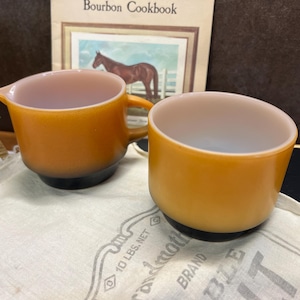 May include: Two vintage amber-coloured glass mugs. One mug has a handle and a spout, while the other is a simple cup. Both have a black base and a light pink interior. The mugs are on a white cloth.