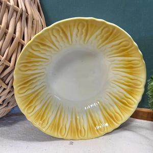 May include: A decorative yellow and white ceramic plate with a textured, sunburst-like pattern radiating from the center. The plate has a scalloped edge and is likely for serving or display.