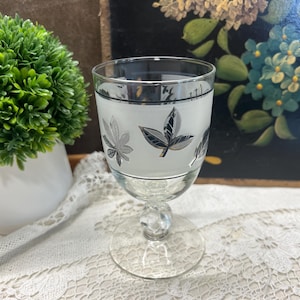 May include: A vintage clear glass goblet with a frosted white band featuring silver leaf designs. The goblet has a clear, round base and a decorative stem. A small green plant in a white pot is in the background.