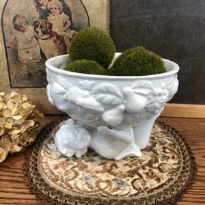 May include: White ceramic bowl with a cherub design and a raised rim. The bowl is filled with green moss balls and is sitting on a brown and cream patterned rug.