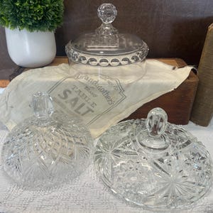 May include: Clear glass lidded jars with intricate cut glass patterns. One jar has a round shape, while the other has a more oval shape. A vintage salt bag and a small book are also in the image.