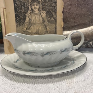May include: White porcelain gravy boat and saucer set. The boat has a curved spout and handle, with a silver trim. The set features a repeating blue and white leaf design. A classic addition to any dining table.