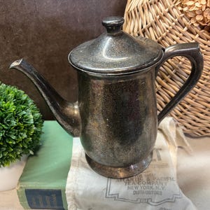 May include: A silver teapot with a tarnished finish and a lid. The teapot is sitting on a white cloth with a label that reads "Atlantic & Pacific Tea Company, New York, N.Y. Distributors".