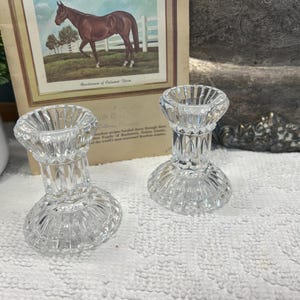 May include: Two clear, cut-glass candle holders with a detailed, geometric design. The candle holders are short and round, with a flared base and a cup-shaped top. A framed print of a horse is in the background.