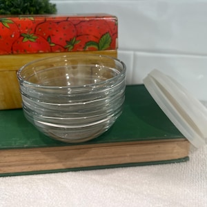 May include: A stack of clear glass bowls, a white silicone lid, and a vintage book with a green cover. A strawberry-themed container is in the background. The bowls are stacked on top of the book.