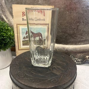 May include: A clear glass tumbler featuring a horse illustration. The glass is placed on a decorative tin. A cookbook with a horse illustration is in the background. The tumbler has a textured design.