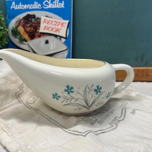 May include: A white ceramic gravy boat with a floral design in teal and gray. The gravy boat has a curved spout and a handle. A recipe book titled "Automatic Skillet Recipe Book" is in the background.