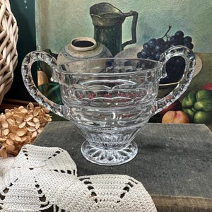 May include: A clear, vintage glass sugar bowl with two handles. The bowl has a decorative pattern and sits on a pedestal base. The background includes a still life painting with fruit and a wicker basket.