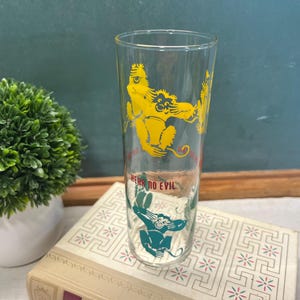 May include: A clear glass with yellow and teal monkey illustrations. The glass has the words "HEAR NO EVIL" in red. The glass is on top of a book with a white and gold design. A small green plant is in the background.