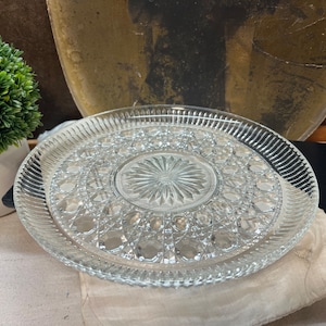 May include: A clear glass serving plate with a raised, decorative design. The plate has a central, starburst-shaped design surrounded by a ring of smaller, raised circles.