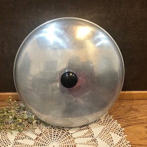 May include: A silver aluminum pot lid with a black knob. The lid is round and has a shiny surface.