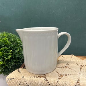 May include: A white ceramic pitcher featuring a vertical ribbed texture along the body and a beaded rim detail. The pitcher has a curved handle on the right side and a small pouring spout on the left side, with a smooth, glossy finish.