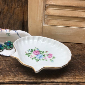 May include: A white porcelain seashell trinket dish with a floral design featuring pink, blue, and white roses. The dish has a gold trim around the edge.