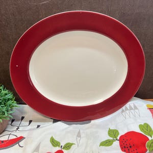 May include: A white oval serving platter with a wide, dark red rim. The platter is on a white tablecloth with a red apple and a yellow and red striped lighthouse design.