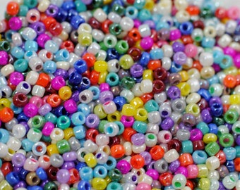 Op de afbeelding: Een grote verzameling kleurrijke zaadkraaltjes in verschillende tinten blauw, groen, rood, geel, paars en wit. De kralen zijn klein en rond, met een lichte iriserende glans.