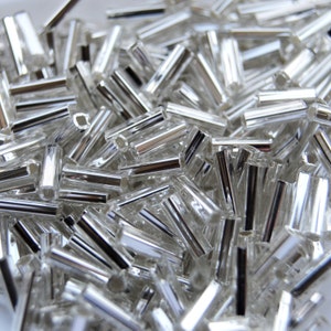 May include: A close-up shot of a pile of clear, cylindrical beads with a silver metallic finish. The beads are scattered, reflecting light and creating a sparkling effect. Ideal for jewelry making and crafting projects.