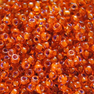 May include: Close-up of a pile of orange seed beads. The beads are small, cylindrical, and translucent, with a purple hue visible in the holes. The beads are scattered, creating a textured surface.