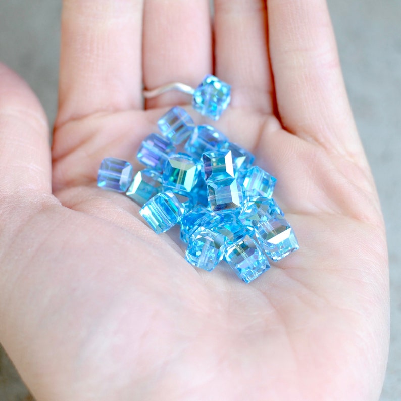 May include: A pile of light blue iridescent square glass beads in a hand.