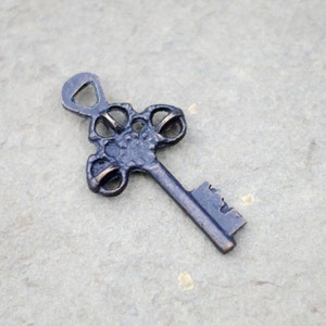 May include: A vintage-style key with an ornate, dark bronze finish. The key features a decorative head with circular cutouts and a detailed key bit. The metal key is likely used for jewelry making, crafting, or as a decorative element.
