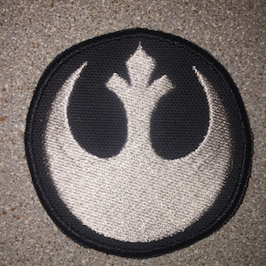 May include: A black and white embroidered patch featuring the Rebel Alliance symbol from Star Wars. The symbol is a white crescent moon with a stylized bird shape in the center.