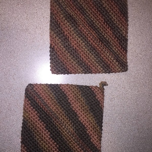 May include: Two square, hand-knitted potholders. The potholders feature diagonal stripes in shades of brown, green, and rust. Each potholder has a small loop in one corner for hanging.