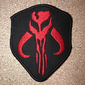 May include: A black embroidered patch with a red Mandalorian symbol. The symbol is a stylized skull with horns and a vertical line down the center.