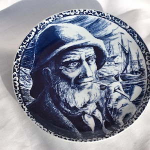 Può includere: Piatto in ceramica blu e bianco con un'illustrazione dettagliata di un uomo barbuto che fuma una pipa. L'uomo indossa un cappello ed è circondato da un bordo di fiori stilizzati.
