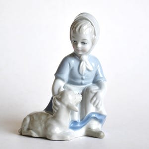 Puede incluir: Figura de porcelana de una figura arrodillada con un atuendo azul claro, con un gorro a juego y un cordero blanco. La figura tiene una expresión serena y es un objeto de colección de temática pastoral.