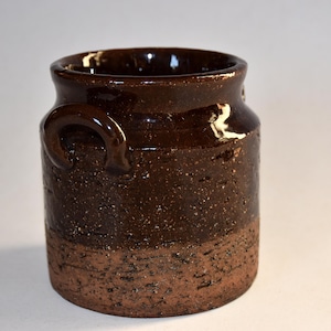 May include: A dark brown ceramic jar with two small handles. The jar has a glossy finish on the top and a textured, rough finish on the bottom. The jar is cylindrical in shape and appears to be handmade.