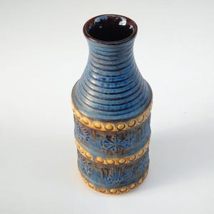 Puede incluir: Un jarrón de cerámica azul con interior marrón y exterior texturizado. El jarrón presenta estrías horizontales cerca de la parte superior y bandas decorativas con patrones en relieve en color marrón claro. El jarrón mide aproximadamente 25 cm de alto.
