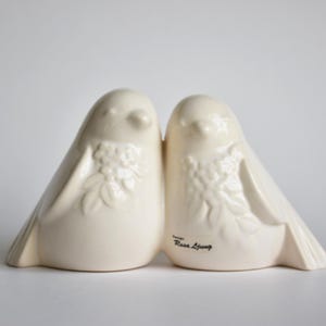 May include: Two off-white ceramic bird ornaments, positioned together. Each bird features a rounded form, small wings, and a floral pattern on the chest. The background is white. The text "Design Rose Living" is visible on one bird.