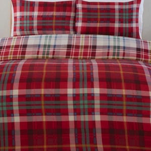 May include: A red tartan bedding set, including two pillowcases and a duvet cover. The pattern incorporates red, white, green, and blue lines. The pillowcases have white edges. The bedding is displayed on a woven headboard.
