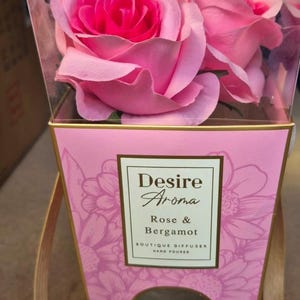 May include: A pink and gold boutique diffuser box with a clear plastic top, containing pink roses. The box features the text "Desire Aroma Rose & Bergamot" and a floral design. A decorative item for the home.