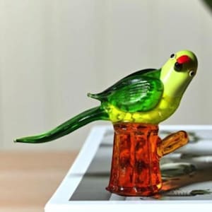 May include: A vibrant glass parrot ornament, perched on a tree stump. The bird has a yellow head, green wings, and a red spot. The tree stump is orange. A decorative item.