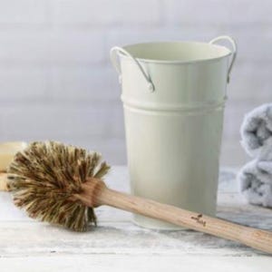 May include: A cream-coloured metal bucket with two small handles, next to a wooden toilet brush with natural bristles. The brush handle is light brown. A folded grey towel is in the background. The items are on a white surface.