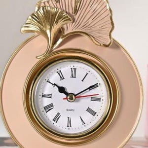 May include: A decorative quartz clock with a blush pink face and a gold-coloured bezel. The clock features black Roman numerals and hands, with a red second hand. The top of the clock is adorned with gold and pink ginkgo leaf accents.