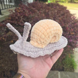 May include: A plush crochet snail toy featuring a light beige body with two upright antennae and a textured, rounded shell in a soft peach color. The yarn has a fuzzy, velvet-like appearance, and the entire piece is crafted with a consistent stitch.