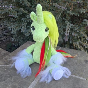 May include: A light green crocheted unicorn plush toy featuring a long, bright yellow mane, a single horn, and large dark eyes. The unicorn has white, fluffy, iridescent-trimmed leg cuffs and a multi-colored tail with red, green, and yellow strands.
