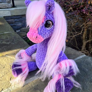 May include: A handmade crochet pony plushie in shades of purple, pink, and white. The pony has a fluffy pink mane and tail, large black eyes, and is sitting on a stone surface. The plushie is a whimsical and cuddly toy.