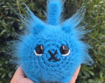 May include: A round, bright blue crocheted plush toy with a fuzzy texture, featuring two large, glossy black eyes with white accents and a stitched black X-shaped mouth. The creature has pointed ears and a small, upright, fuzzy blue tail.