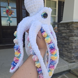May include: A handmade white crochet octopus plush toy with rainbow pom-poms along its tentacles. The octopus has a large head and big, colorful eyes. The plush is soft and fluffy, perfect for a child's toy or nursery decor.