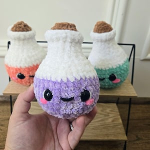 May include: Three plush potion bottles in orange, purple, and green, each with a white top and a brown cork. The bottles have smiling faces with pink cheeks and black eyes. The purple bottle is held in a hand.